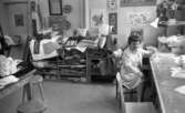 Västra mark 5 juni 1968

I textilverkstaden på Västra marks sjukhus sitter en kvinna och dricker kaffe vid ett högt bord i förgrunden. Hon är klädd i en vit kofta och vitrandig klänning. Hon har en vit ring på höger ringfinger. En annan kvinna sitter delvis skymd på en stol bakom henne. En massa dukar och kuddar m.m. ligger på hyllor i bakgrunden.