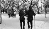 Ånnaboda 29 januari 1968

Två män med skidor över axlarna och stavar i händerna går på en skogsväg i Ånnaboda. De är klädda i svarta jackor, svarta byxor och svarta pjäxor och står med ryggarna mot kameran. Mannen till vänster har en ljus mössa med tofs och mannen till höger har en svart mössa med tofs. I bakgrunden syns andra personer.