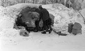 Ånnaboda, fotojobb (?), arbetarkommun 70 år 12 februari 1968

En kvinna och två tonårspojkar befinner sig ute i naturen i Ånnaboda. De befinner sig vid sidan om en snödriva med en utskjutande kant över. Det ser ungefär ut som en halv grotta. De har tak över sina huvuden. Kvinnan sitter ned och de unga tonårspojkarna står upp. Kvinnan är i färd med att ge en av pojkarna skräp som han lägger i en plastpåse. Alla tre är klädda i mössor, jackor, byxor och pjäxor. En annan person sitter delvis skymd i bakgrunden. Till höger ligger en liten pulka samt tre lådor på marken.