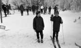 Ånnaboda, fotojobb (?), arbetarkommun 70 år 12 februari 1968

Två pojkar i tioårsåldern står i skidspåren i Ånnaboda. Pojken till höger har stavar i händerna och pjäxor samt skidor på fötterna. Båda pojkarna är klädda i mörka mössor, mörka jackor, mörka byxor och ljusa vantar. Pojken till vänster har också skidor på fötterna. Många personer syns i bakgrunden.