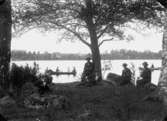 Sjöutsikt, grupp vid sjön, några personer i roddbåtar.
Bostadshus i bakgrunden.