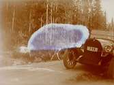 Bil vid skogsväg. En get går utmed vägen. 
Bilen är en Oldsmobile modell 30 från 1923-24 med en 42 hästkrafters sexcylindrig motor. Den registrerades den 2:e april 1924 på fröken Jenny Larsson, Örebro, och fick registreringsnummer T99. År 1930 ägdes den fortfarande av Jenny Larsson.
