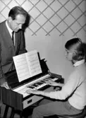 Rumsinteriör, en flicka vid pianot, en man bredvid.
Skandia Musik