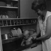 Rumsinteriör, en liten pojke som badar i diskhon, en kvinna.
Patrik och Plutt