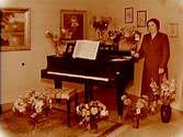Fru Falk, pianolärarinna vid ett piano.
Rumsinteriör, högtid.