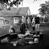 Familjegrupp fyra personer vid bilen. Bostadshus i bakgrunden.
Hallebys resa.