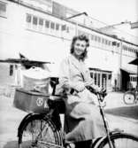 Konsums springflickor, en flicka med cykel.
Affärsbyggnad i bakgrunden.