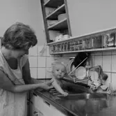 Rumsinteriör, en liten pojke som badar i diskhon, en kvinna.
Patrik och Plutt