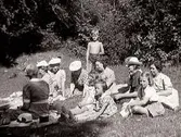 Utflykt. Kvinnor och några barn på utflykt. Sällskaper sitter på en äng med träd i bakgrunden.