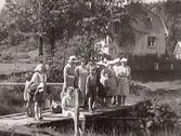 Utflykt. Kvinnor och några barn på utflykt. Sällskapet står på en liten bro. I bakgrunden syns ett bostadshus.