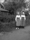 Två kvinnor i folkdräkt.
Bostadshus i bakgrunden.
Dalarna