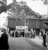 Festligheter vid Siggebohyttan den 1 augusti 1937. Folk är samlade på gårdsplanen vid Siggebohyttan. Vid västra delen av Siggebohyttans bergsmansgård pågår ett tombolalotteri. I bergsmansgårdens svalgång står folk och tittar ut genom fönstren.