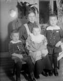 Rumsinteriör, familjegrupp fyra personer.
Edit Andersson med barnen (från vänster) Lars (givaren), Britta och Olle. Bilden är tagen i hemmet i Järnhandelshuset i Norberg där familjen bodde från 1912. 1926 flyttade de därifrån till huset Margretehill i Norberg. Bilden kan vara tagen 1917 då Britta är ca 1 år.
