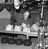 Lindbergs Strå- och Filth. Hattillverkning. 216/67 Stumpar på spassfåglar hänger i taket, fyra svarta hattar utan brätten så kallad mössor i förgrunden. I bildens centrum olika träformar. I förgrunden till höger tvingar.