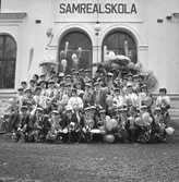 Samrealskola, gruppfoto, studenter.
Skolbyggnad i bakgrunden.