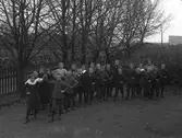Skolfoto. Skolklass fotograferad utomhus, utanför folkskolan i Karl Gustav. Barnen är uppställda parvis, som till dans. Lärarinnan heter Anna Modin.