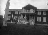 Tvåvånings bostadshus.
Grupp framför huset.