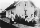 Släktgrupp åtta personer, kaffebjudning.
Bostadshus i bakgrunden.
Från höger: Erik Theon Olsson, sonen Sven Erik, frun Emma Lydia Olsson, fru Olssons föräldrar Fredrik och Johanna Andersson, Olssons döttrar Linnéa och Thea Elfrida och sist till vänster Alma Andersson, fru Olssons syster.