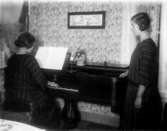 Rumsinteriör, två flickor vid pianot.
Erik Theon Olssons döttrar, Thea Elfrida och Linnéa prövar taffeln.
Bilden tagen 1928 eller 1929.