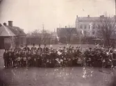 Elementarskolan i Trelleborg, firandet av Gustaf Adolfsfesten 1894, Södra skolgården med rektor, lärare och elever, foto fr. N mot S. den (6nov 1894?) i fonden hamnen. Skolgården begränsas av ett träplank, med portgrind. Till höger Rektor Sjövalls hönshus vid planket, därintill ett boketräd(Fagus) Husen: från vänster 1: Skolans hemlighus (under skiffertak) sammanbyggt med 2: ”Rektor Sjövalls tvättstuga”, m duvslag på taket. 3: ”Skräddare Ant. Nilssons hus” 1-vån. med 1st. skorsten, bel. Å tomt nr. 19 (59B) inom kv. Herkules. 4: ”Tryckarehusen” 2 våningsbyggn. Uppf. Av boktryckarna Sandberg och Jönsson å tomt nr 20 (61B) inom kv. Herkules. Äldre personer från vänster: 1: Rektor Hjalmar Sjövall, fil dr 2: Kollega Elof ,fil dr 3: Kollega John Smith, fil dr 4: Kollega Alexander Rydberg, pastor
5: Flickskolelärarinnan fröken Charlotte Jönsson i spetssammetshatt (nackhatt)    6:Lärarinnan fröken Mimmi Rosqvist i bred hatt med sidenband 7: Lärarinnan Emmy Ohklander dotter av Framför det förstnämnda huset, synas den med halm vinterinklädda träpumpen
5146 Axel Andersson.
