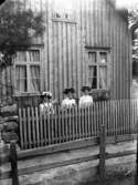 Gruppbild av tre kvinnor fotograferade utomhus stående vid en husvägg bakom ett staket.