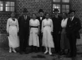 Fyra män och fyra kvinnor stående vid en husvägg utomhus, 1919
	Metallutfällning.