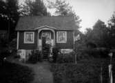 Liten stuga, två kvinnor en man och ett barn framför, ”1915 B” på asken
	Metallutfällning.