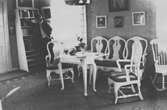 John Bauer står vid bokhyllan i ett rum på Björkudden. Mellan fönstren är en soffa och två stolar placerade runt ett bord.