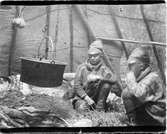 John Bauers Lapplandsresa sommaren 1904. I en kåta sitter två män vid eldstaden där en gryta hänger i en kedja från taket.