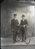 Gruppbild, två (2) män stående i helfigur. Ateljébild. Den ena mannen håller ytterkläder över ena armen och bär keps. Andra mannen har plommonstop och lutar sig mot cykeln. Beställare: Axel Andersson.
