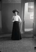 Kvinnoporträtt, kvinna stående i helfigur iförd hatt. Ateljébild. Beställare: Alma Andersson, Fageråkra.