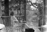 På Björkuddens veranda sitter Ester Bauer i en gungstol med tidningen i knät och vid hennes fötter ligger hunden. En stig går genom skogen ner mot sjön.