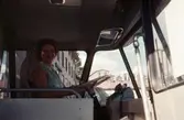 En kvinnlig busschaufför i sin buss vid Gamla Torget i Mölndal, 1970-tal. I bakgrunden, genom bussens rutor, skymtas husen från vänster Kvarnbygatan 41, 43 samt taket på nr 45.

För mer information om bilden se under tilläggsinformation.