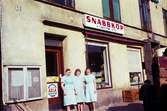 Tre kvinnliga butiksbiträden fotograferade utanför snabbköp med adress Kvarnbygatan 43 vid Gamla Torget i Mölndal, 1960-tal.

För mer information om bilden se under tilläggsinformation.