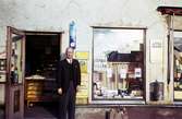 En man ståendes i dörren till tobakshandeln med adress Kvarnbygatan 43 vid Gamla Torget i Mölndal, 1960-tal.

För mer information om bilden se under tilläggsinformation.