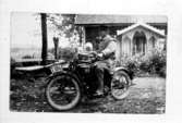 Fotografi från Södra Billinge, Kils socken. Året är 1920. Hugo Hammarlund har inköpt en motorcykel  Excelsior Chicago, 1000 cc, 1920 års modell för 3000:-.
I sidovagnen sitter brodern Gustaf med sin dotter Ingrid, född år 1919. I bakgrunden deras föräldrahem i Södra Billunge by. Observera den vackra snickarglädjen på verandan.