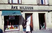 Två kvinnor står utanför dörren till Åke Nilsson charkuteri och delikatesser med adress Kvarnbygatan 43 vid Gamla Torget i Mölndal, 1960-1970-tal.

För mer information om bilden se under tilläggsinformation.