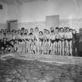 En samling med barn står på kanten av bassängen i Huskvarnas badhus. Till vänster sittande på huk simlärare Erik-Arne 
