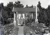 En man och en kvinna vid en liten stuga i Kalmar södra koloniområde, fotograferat omkring 1930. Kalmar södra koloniförening grundades 1917 och har idag 105 kolonilotter. Området ligger strax söder om länssjukhuset i Kalmar med huvudingång från Stensbergsvägen.