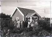 En stuga och en grupp människor i Kalmar södra koloniområde, fotograferat omkring 1930. Kalmar södra koloniförening grundades 1917 och har idag 105 kolonilotter. Området ligger strax söder om länssjukhuset i Kalmar med huvudingång från Stensbergsvägen.