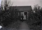 En stuga, man och kvinna i Kalmar södra koloniområde, fotograferat omkring 1930. Kalmar södra koloniförening grundades 1917 och har idag 105 kolonilotter. Området ligger strax söder om länssjukhuset i Kalmar med huvudingång från Stensbergsvägen.