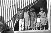 Fem barn, pojkar och flickor, står framför en randig tältduk. De håller varandra om axlarna. En av pojkarna har en cigarrett i munnen. Familjen Taikons läger i Johanneshov, södra Stockholm.