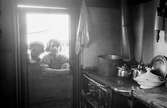 Interiör från familjen Taikons läger vid Johanneshov, södra Stockholm. På köksspisen står kastrull, kaffekannor och en terrin. Två barn kikar in genom dörrens öppna övre halva.