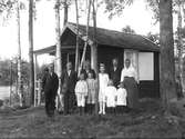 En grupp vuxna och barn framför en liten stuga i skogen.