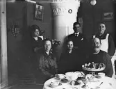 Sex personer vid kaffebord. Kakelugn i bakgrunden.
