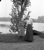 Kvinna och flicka vid stranden. Bilden tagen mot nedre dammen och tegelmagasinet i Gammelstilla, Torsåker.