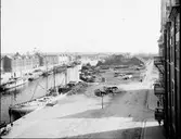 En bild från en levande hamn med fartyg, varv, liv och rörelse. Dalapalatset till höger. På Hellbergska huset vajar flaggan på halvstång.

