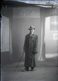 Mansporträtt, man stående i helfigur. Ateljébild. Iförd hatt och lång ytterrock, samt håller spatserkäpp i höger hand. Beställare: John Andersson, Sällstorp.
