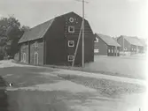 Gamla magasinet invid infarten. Till höger ladugården timmer i korsknut. Brutet tak. Huset rött med vita foder.