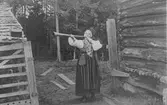 En kvinna i folkdräkt, som blåser i en näverlur, vid en fäbod.